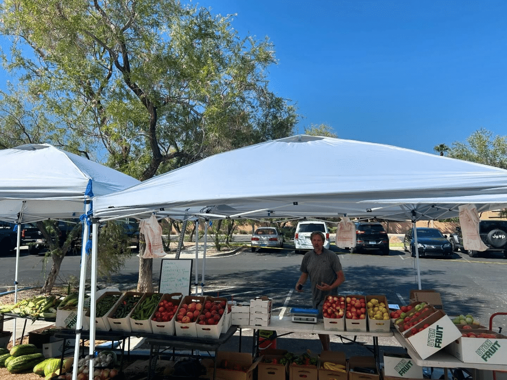 Man under two tents with fruit and vegetables in cardboard boxes that say "Utah Fruit" at Fresh 52 Farmers Market